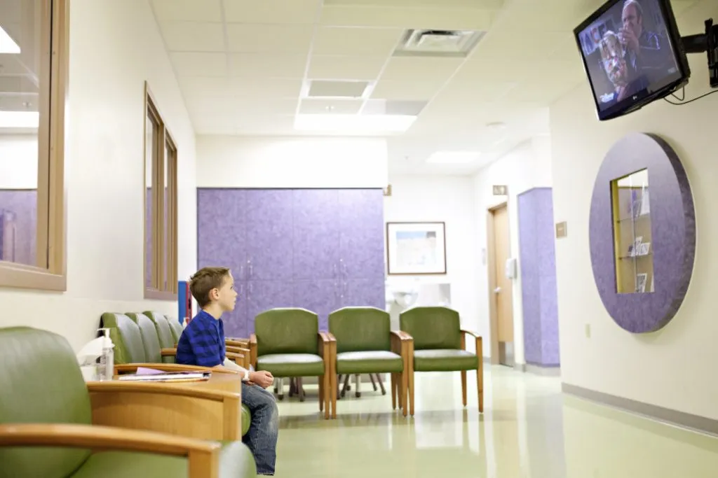 A boy sits in a waiting lobby at a doctor's office watching a television.