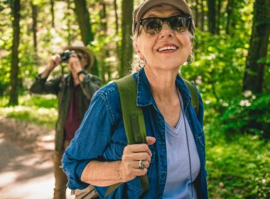 A woman hiking through the forest in the spring while her partner uses binoculars to bird watch behind her.